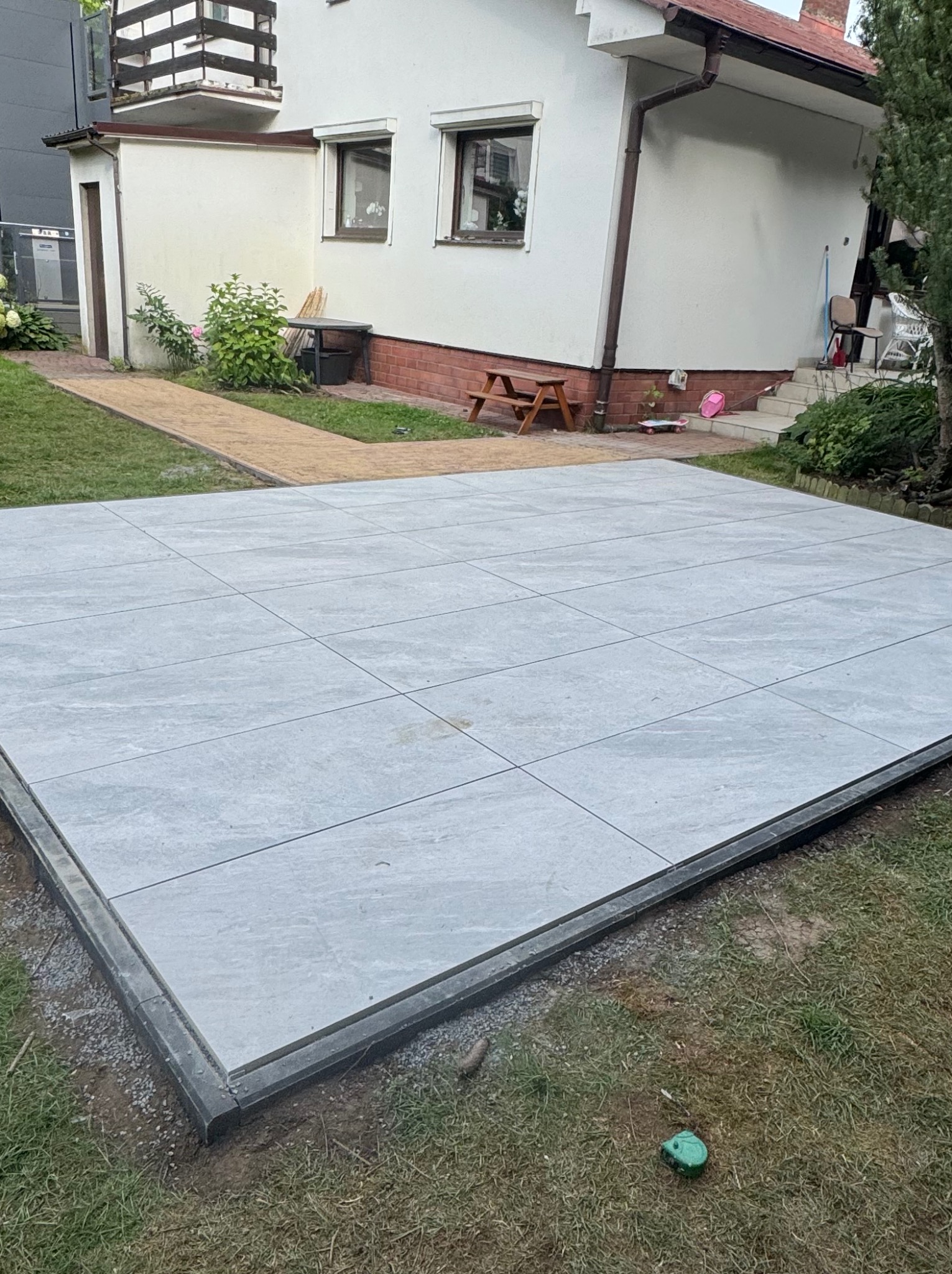 Jasnoszara, kwadratowa nawierzchnia z płyt ułożona na trawie, obrzeża z ciemnoszarego betonu. W tle dom z beżową elewacją i brązowymi elementami wykończeniowymi.