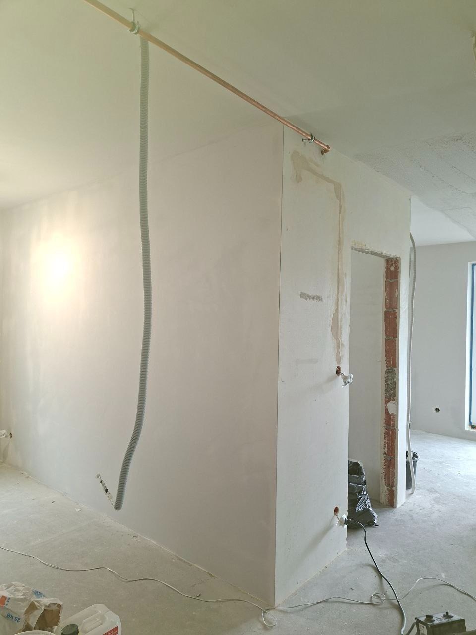 Wnętrze po szpachlowaniu ścian, widoczne gładzie, rurka miedziana pod sufitem i peszel. Na podłodze wiadro z farbą. Ściany przygotowane do malowania.