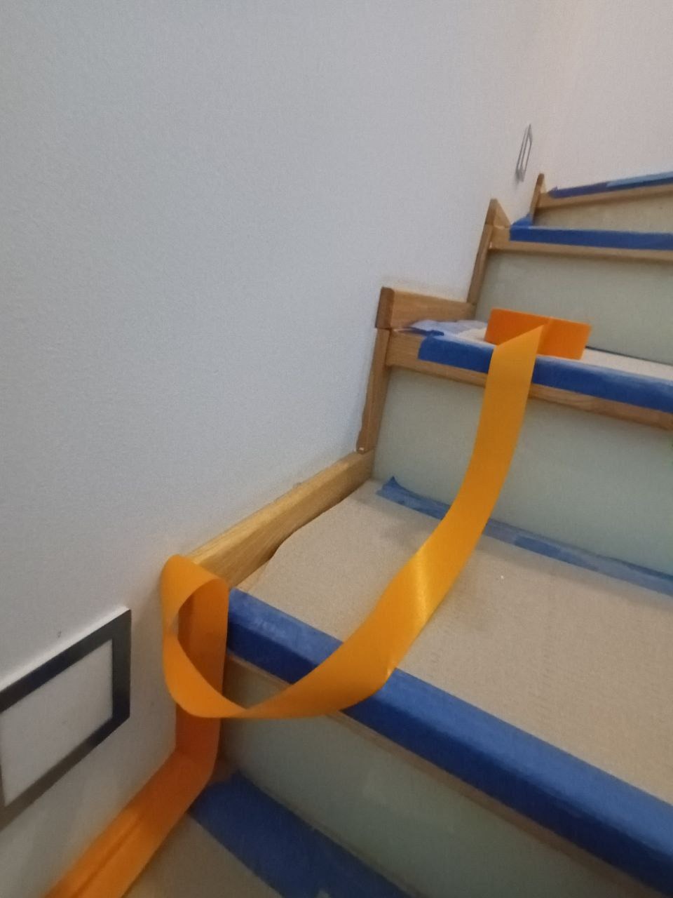 Schody zabezpieczone niebieską taśmą malarską i pomarańczową folią ochronną przed malowaniem. Widoczny fragment ściany z gniazdkiem.