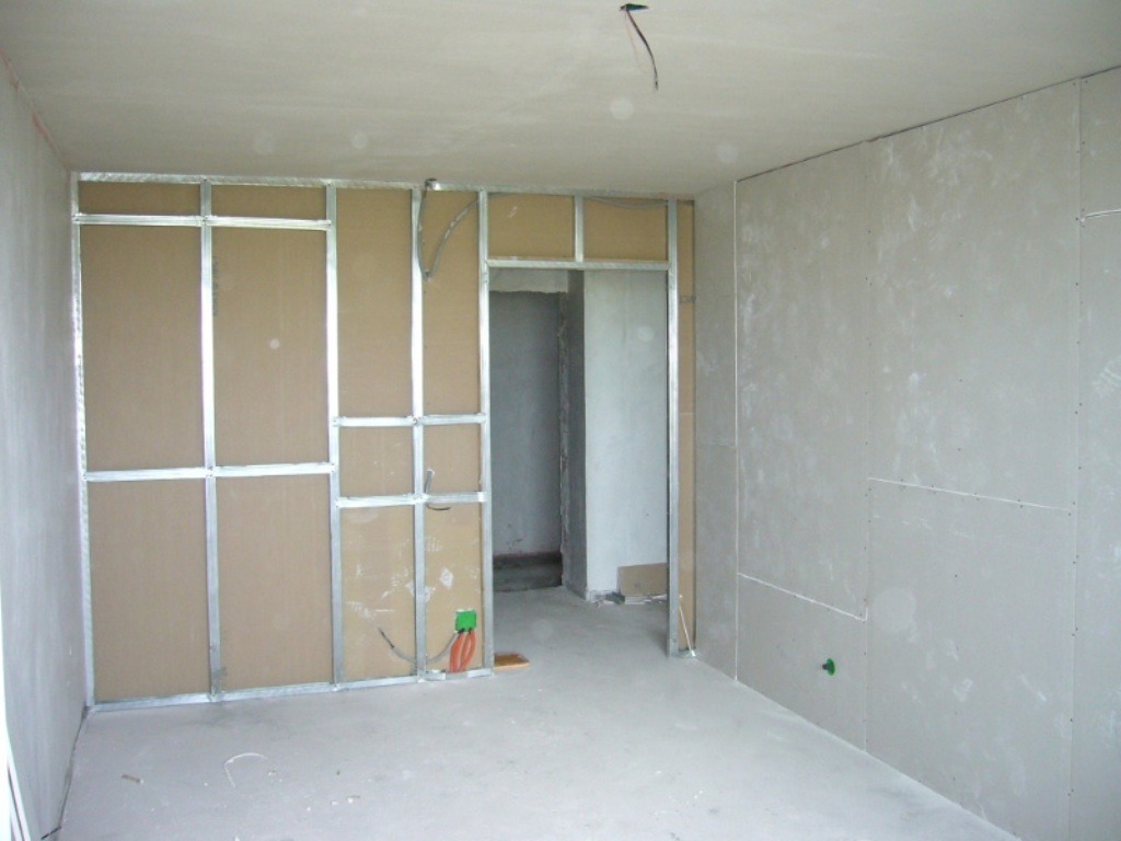 Montaż ścian działowych z płyt karton-gips na metalowym stelażu w surowym wnętrzu. Widoczne przewody elektryczne i otwór drzwiowy.