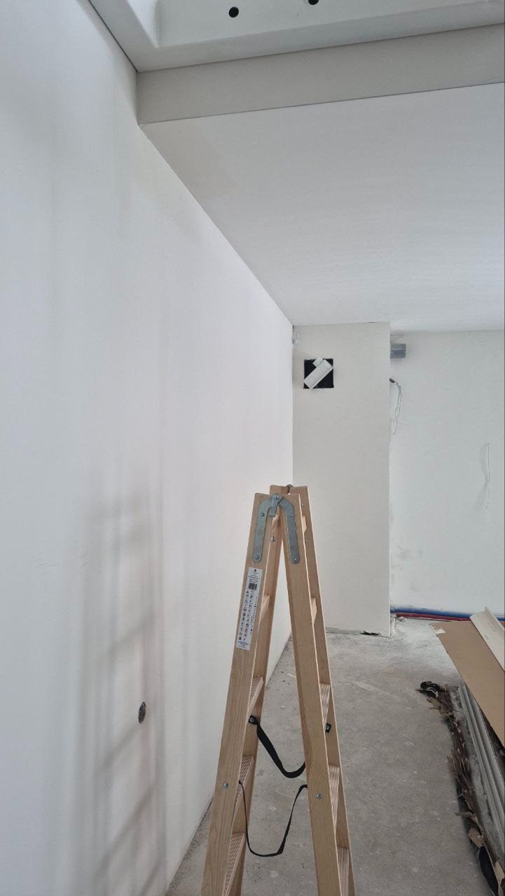 Wnętrze po szpachlowaniu: gładka, biała ściana z widoczną drabiną. Przygotowanie do malowania, widoczne elementy instalacji i wykończenia sufitu.