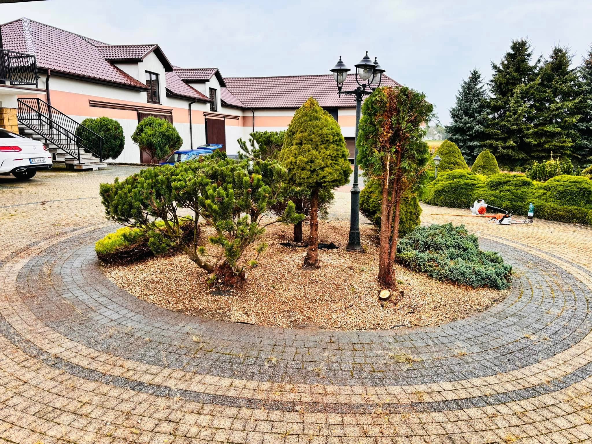 Ogród z okrągłą rabatą, niskie drzewa i krzewy, kostka brukowa. W tle dom z beżową elewacją i ciemną dachówką. Widoczna piła spalinowa.