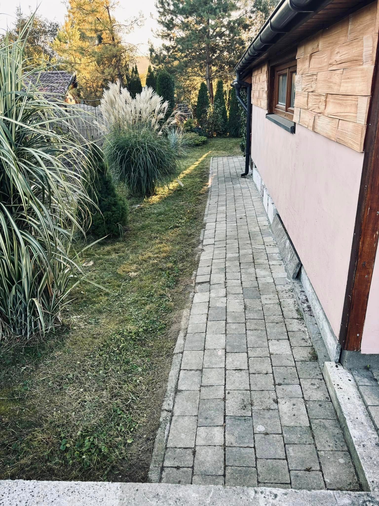 Ogród z ozdobnymi trawami i ścieżką z kostki brukowej wzdłuż budynku z drewnianą elewacją. Jesienne słońce oświetla roślinność.