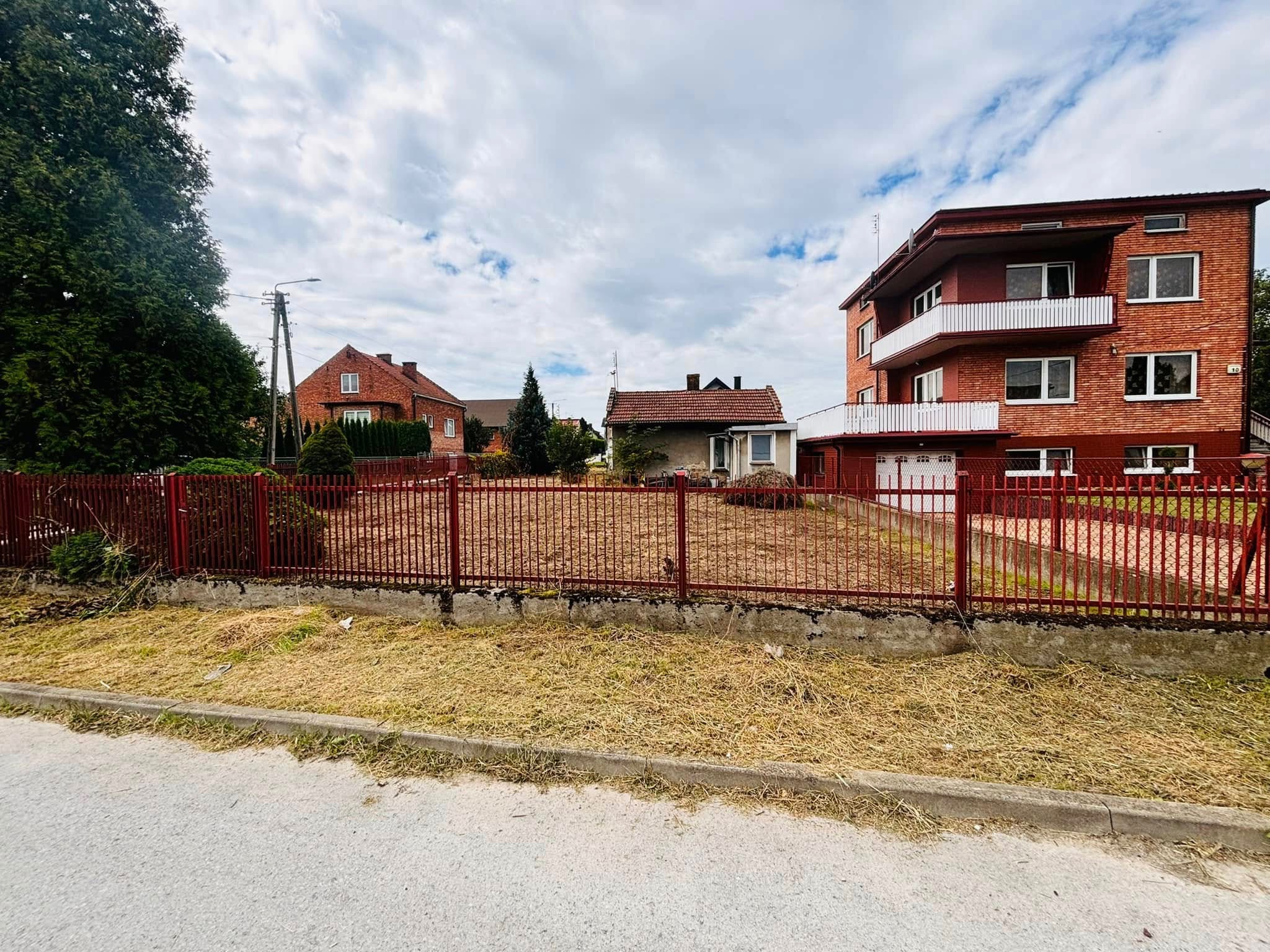 Działka z rudą ziemią, otoczona czerwonym metalowym płotem, z widokiem na domy w tle. Świeżo skoszona trawa przy chodniku.