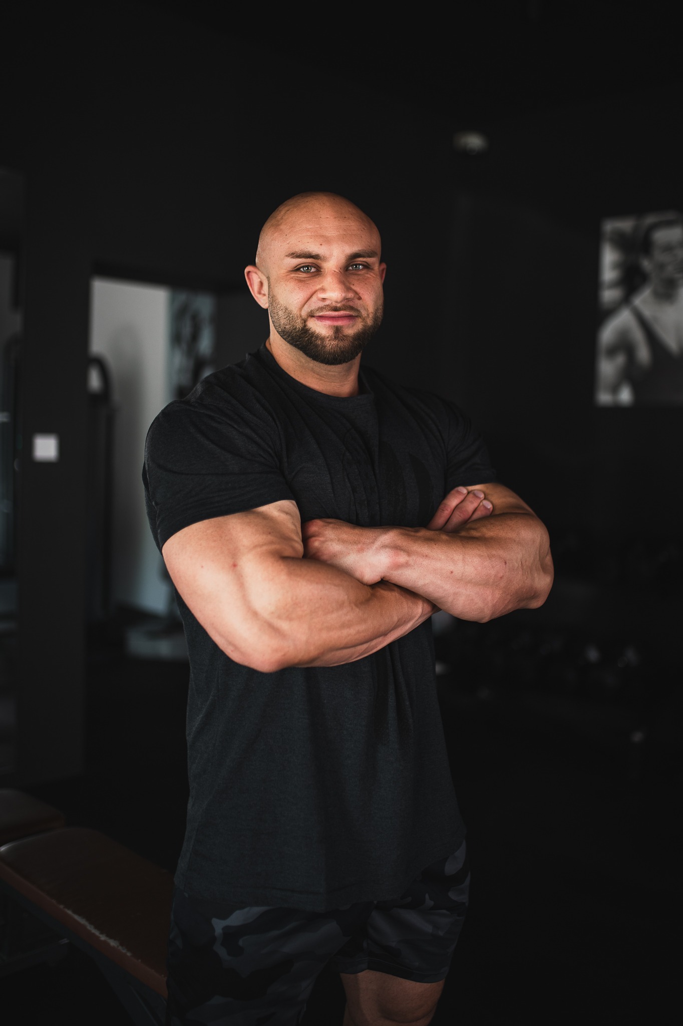 Uśmiechnięty mężczyzna z brodą, w czarnej koszulce, stoi na tle siłowni z założonymi rękami, prezentując muskulaturę.