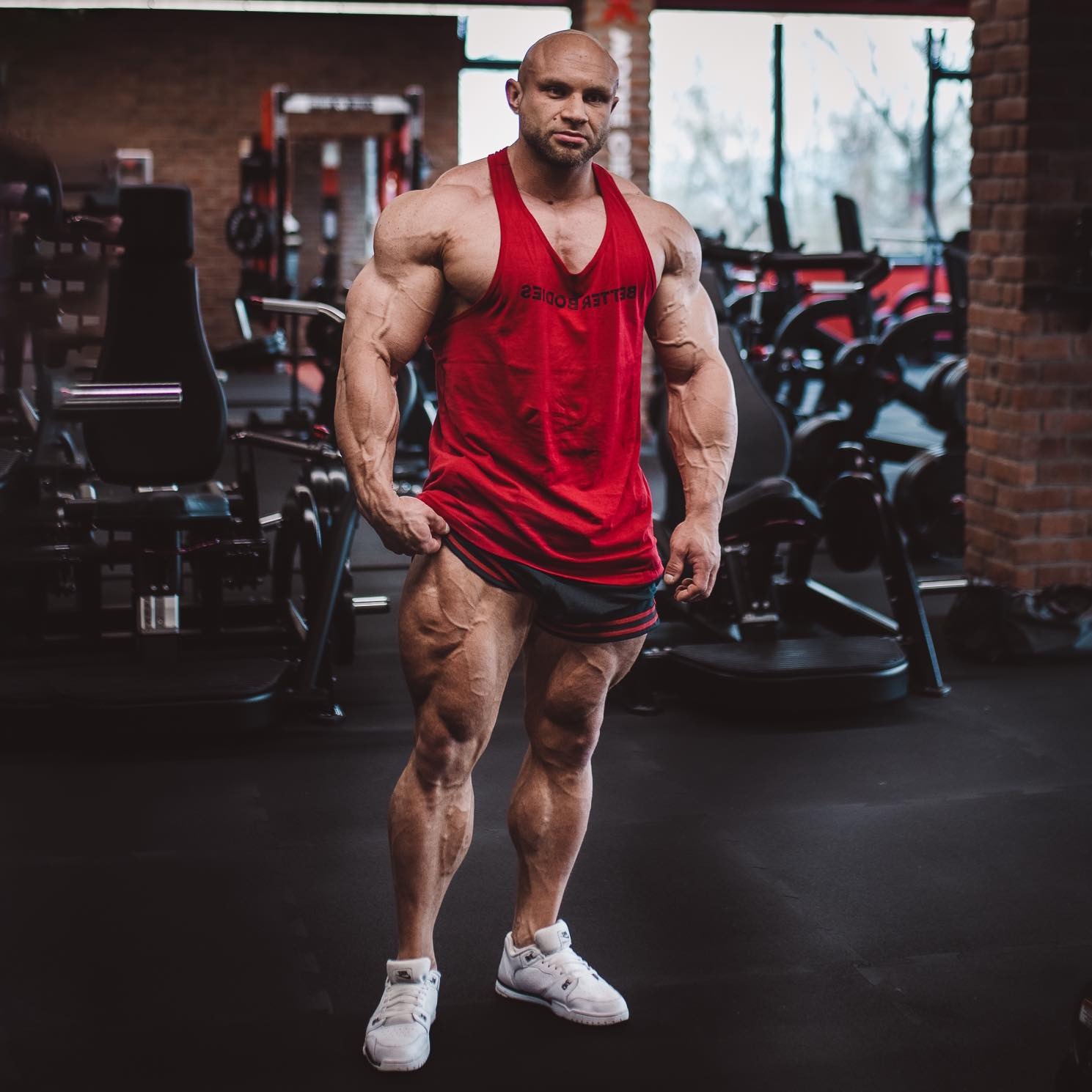Umięśniony mężczyzna w czerwonej koszulce pozuje w siłowni, prezentując imponującą muskulaturę nóg i ramion. W tle sprzęt do ćwiczeń i ceglana ściana.
