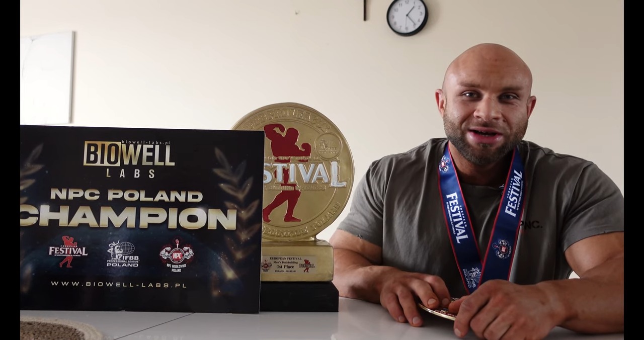 Uśmiechnięty mężczyzna z medalem na szyi, obok nagroda i plakat Biowell Labs NPC Poland Champion. Zegar na ścianie w tle. Prezentacja osiągnięć sportowych.
