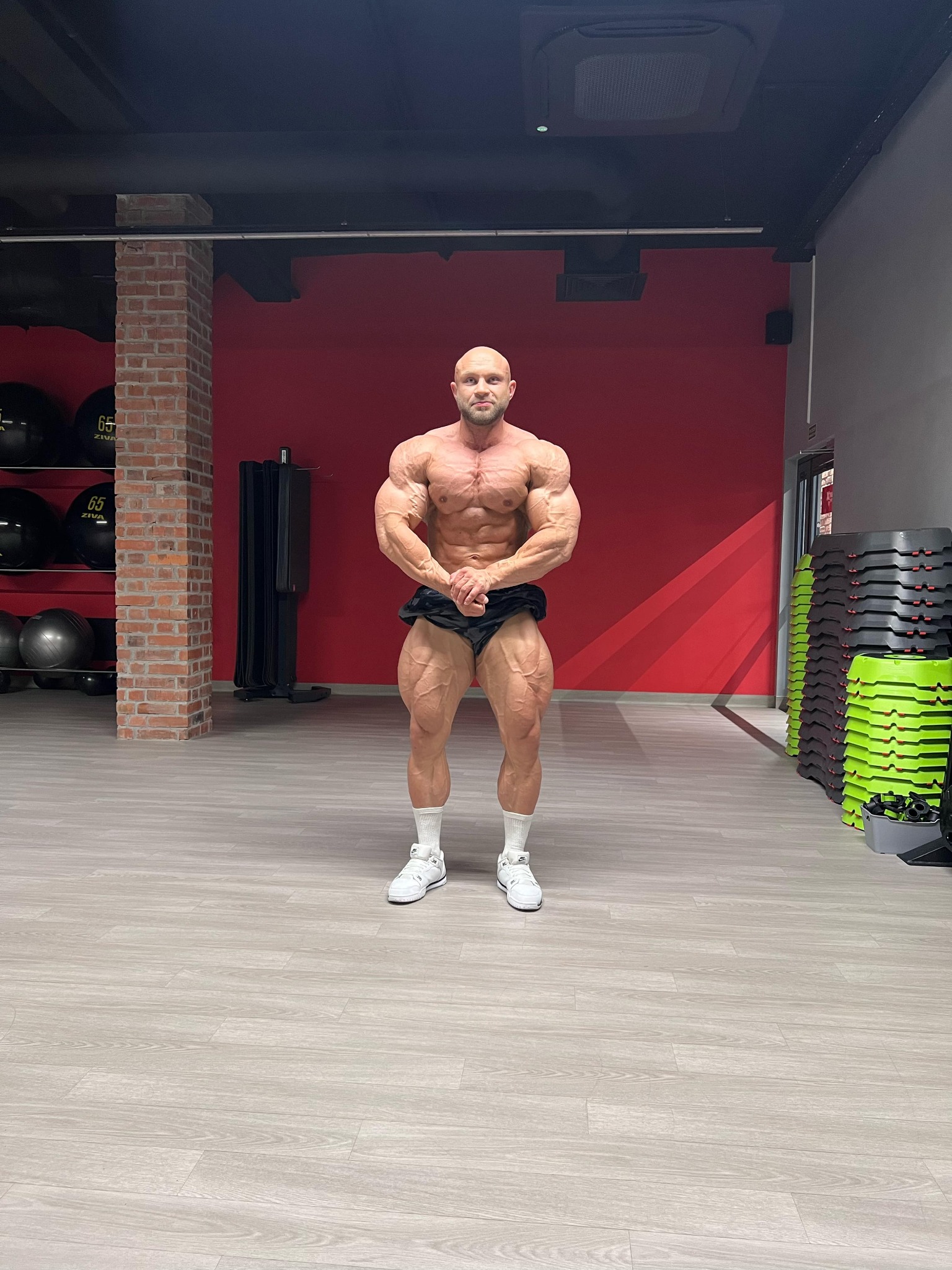 Umięśniony mężczyzna w sportowych szortach, białych skarpetkach i butach, prezentuje sylwetkę na tle czerwonej ściany w siłowni. Widoczne piłki i stepy.