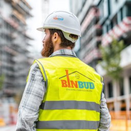 GrafIT Center Daniel Jurek - Pracownik w Radoszycach w kamizelce z logo Binbud i kasku z logo, na tle budowy. Prezentacja projektu logo na odzieży roboczej.