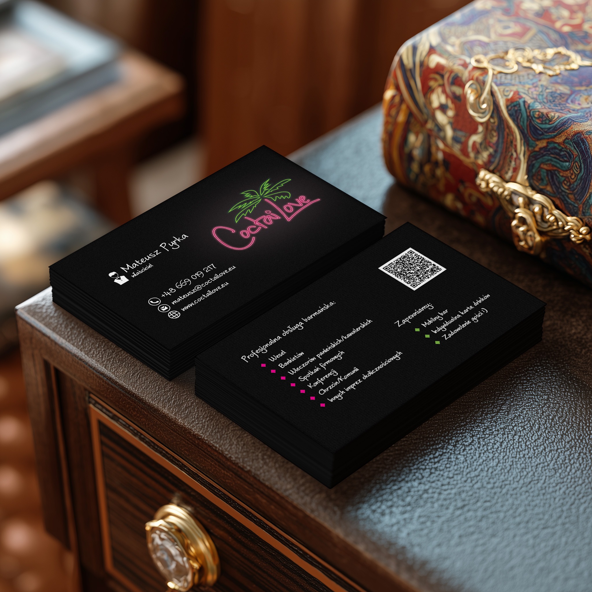 Stos czarnych wizytówek CoctaiLove z neonowym logo na drewnianej komodzie, obok zdobiona szkatułka. Elegancki projekt wizytówki z danymi kontaktowymi i QR kodem.