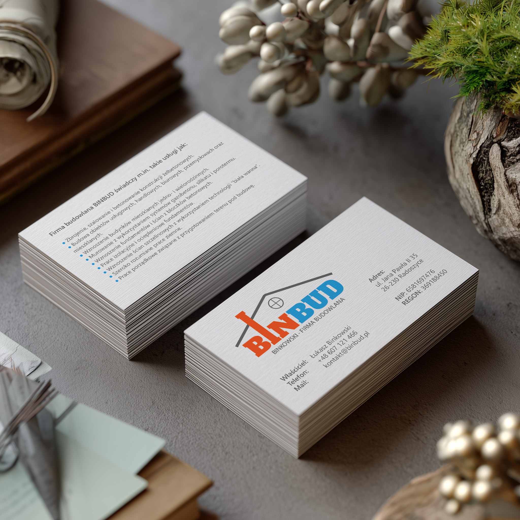 Eleganckie wizytówki BinBud z logo w kolorach pomarańczowym i niebieskim, na teksturowanym tle. Widoczne dane adresowe i kontaktowe. Minimalistyczny design, profesjonalny wygląd.