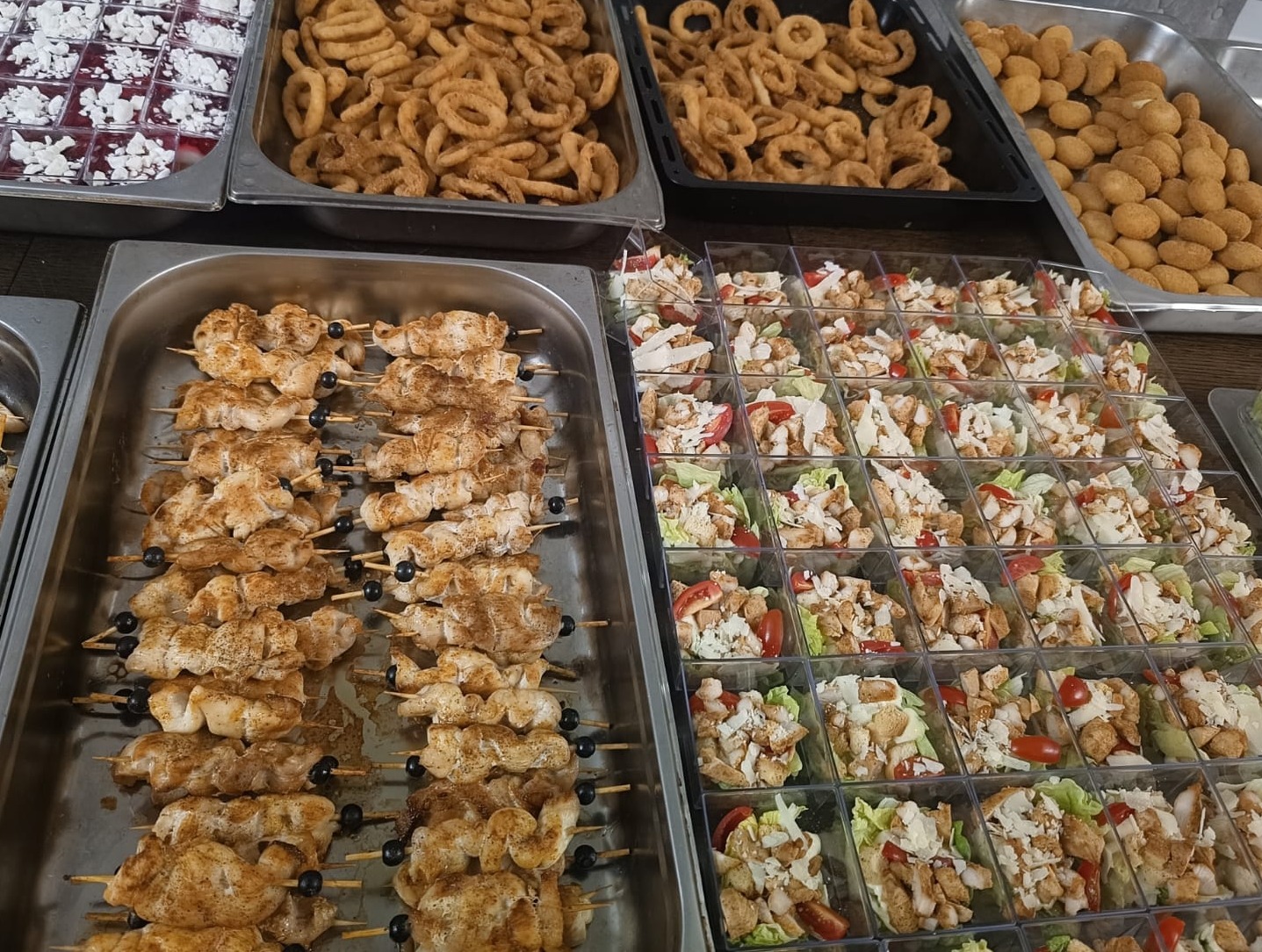 Różnorodny catering: szaszłyki z kurczaka, sałatki w kubeczkach, krążki cebulowe i kulki serowe na metalowych tacach, gotowe do serwowania.