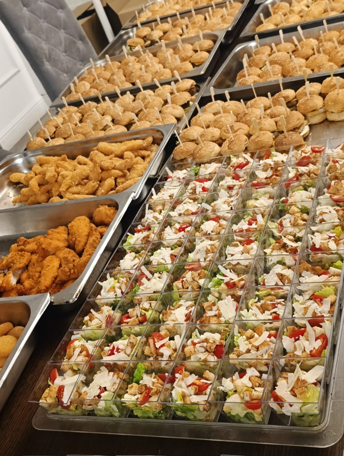 Bogaty wybór przekąsek cateringowych: mini burgery z wykałaczkami, smażone kąski kurczaka i sałatki w przezroczystych pojemnikach, ułożone na metalowych tacach.