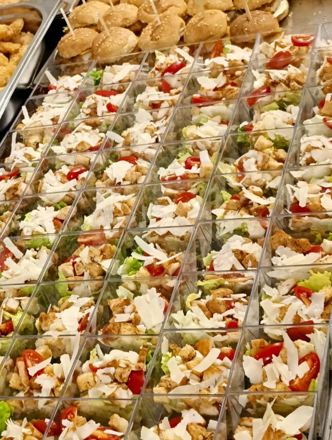 Mini porcje sałatek z kurczakiem i pomidorami w przezroczystych pojemnikach, obok mini burgery na tacy. Gotowe dania cateringowe.