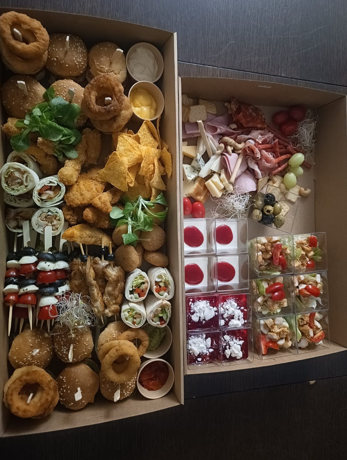 Dwie kartonowe tace z różnorodnymi przekąskami: mini burgery, tortille, koreczki, nachosy, sery, wędliny, sałatki w kostkach i desery w przezroczystych pojemnikach.