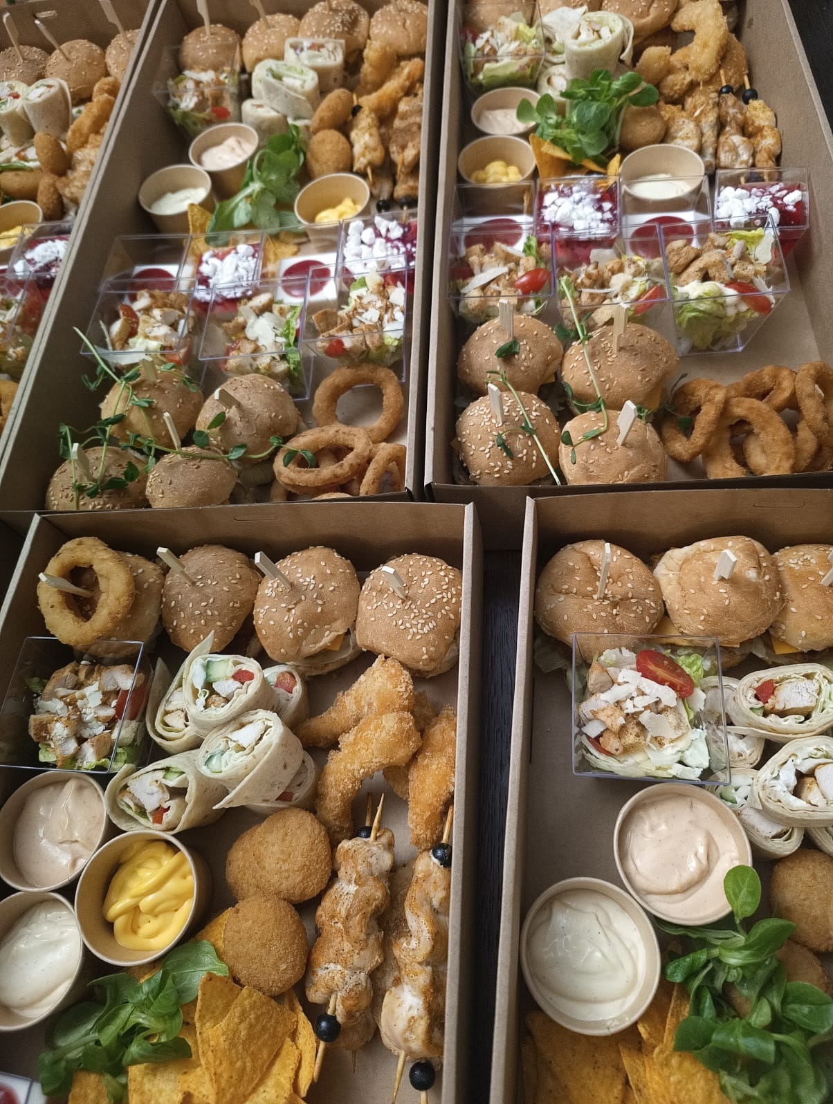Zestaw różnorodnych przekąsek cateringowych w kartonowych pudełkach: mini burgery, tortille, sałatki w kubeczkach, krążki cebulowe, nuggetsy i sosy. Widok z góry.