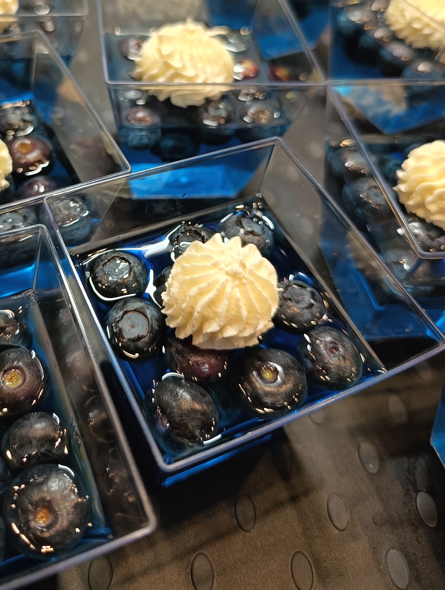 Deser w przezroczystym pojemniku: borówki w niebieskiej galaretce, zwieńczone kremem. Widok z góry, ostre światło podkreśla detale.