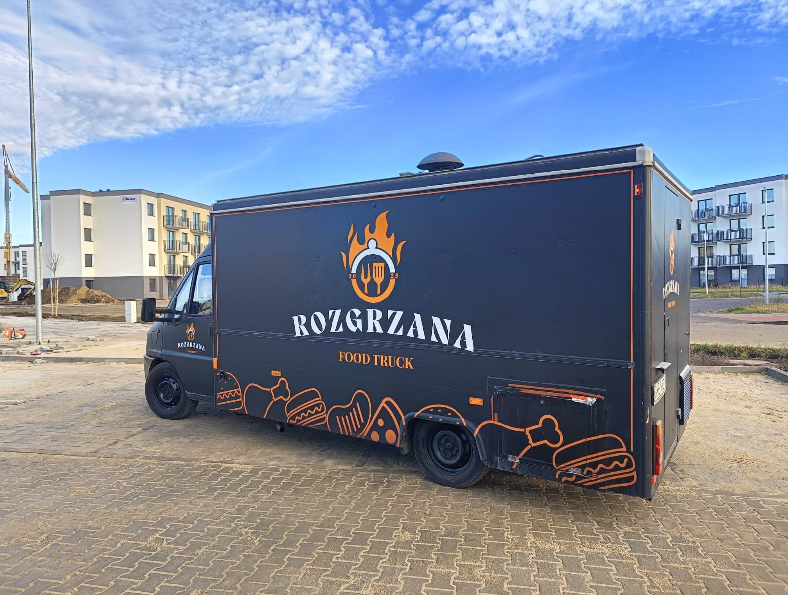 Czarny food truck 'Rozgrzana' z pomarańczowymi grafikami jedzenia na tle błękitnego nieba i budynków w trakcie budowy. Widok z boku, lekko z dołu.