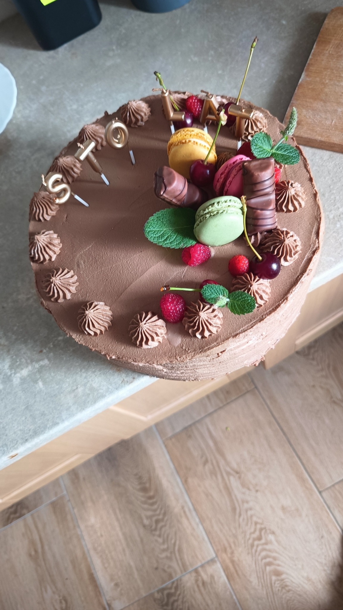 Czekoladowy tort urodzinowy z makaronikami, malinami, wiśniami i świeczkami układającymi się w napis 'STO LAT', sfotografowany z góry na blacie kuchennym.