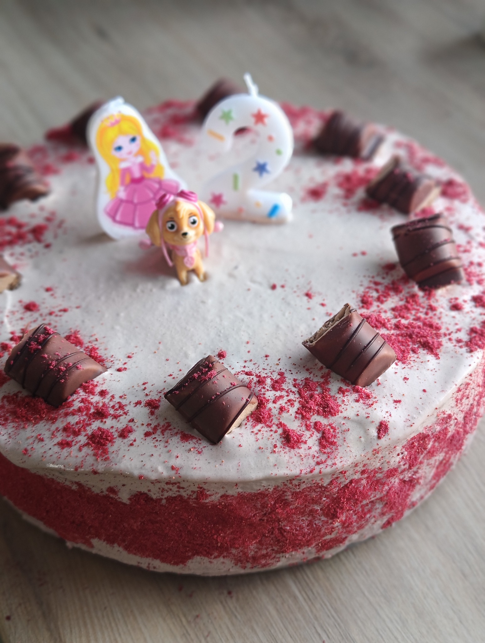 Tort urodzinowy z kremem, posypką malinową, dekoracjami z czekolady, świeczką z postacią z bajki i figurką pieska.