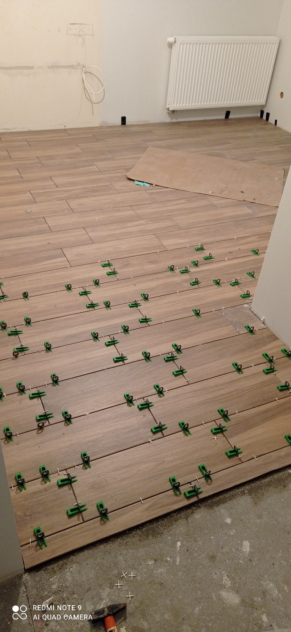Układanie podłogi z płytek imitujących drewno, z użyciem zielonych klipsów poziomujących. Widoczny fragment grzejnika i wystające przewody elektryczne.