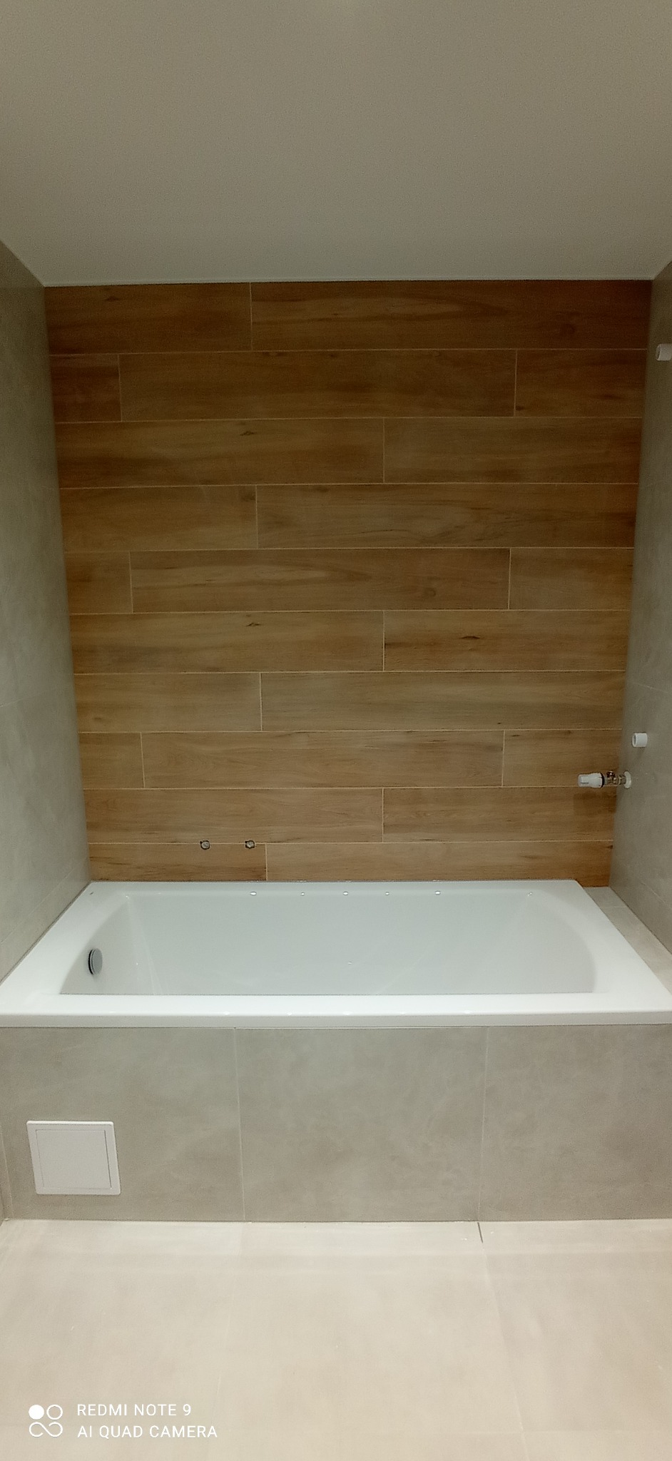 Wykończona łazienka z wanną obudowaną płytkami w odcieniach beżu i brązu. Na ścianie płytki imitujące drewno. Minimalistyczny styl, widok z przodu.