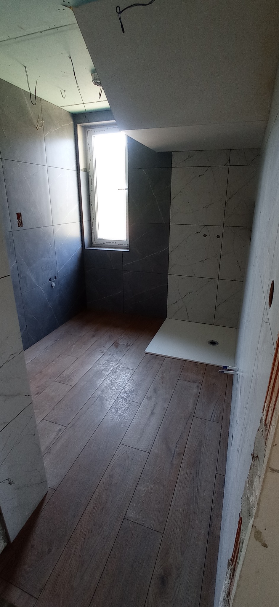 Wnętrze łazienki w trakcie remontu: ułożone płytki podłogowe imitujące drewno, częściowo wykończone ściany płytkami w odcieniach szarości i bieli, widoczne okno.