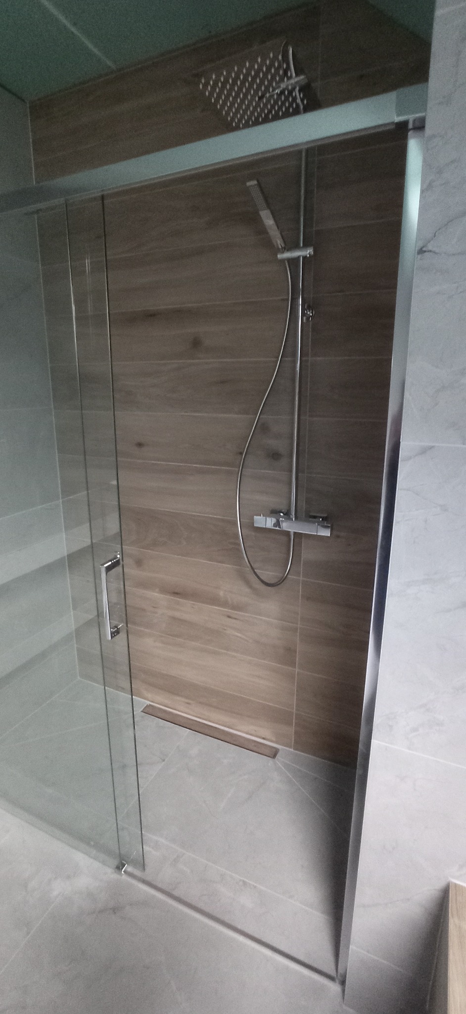 Nowoczesna kabina prysznicowa z przesuwnymi drzwiami, wykończona płytkami imitującymi drewno i szarym marmurem. Widoczny odpływ liniowy i deszczownica.