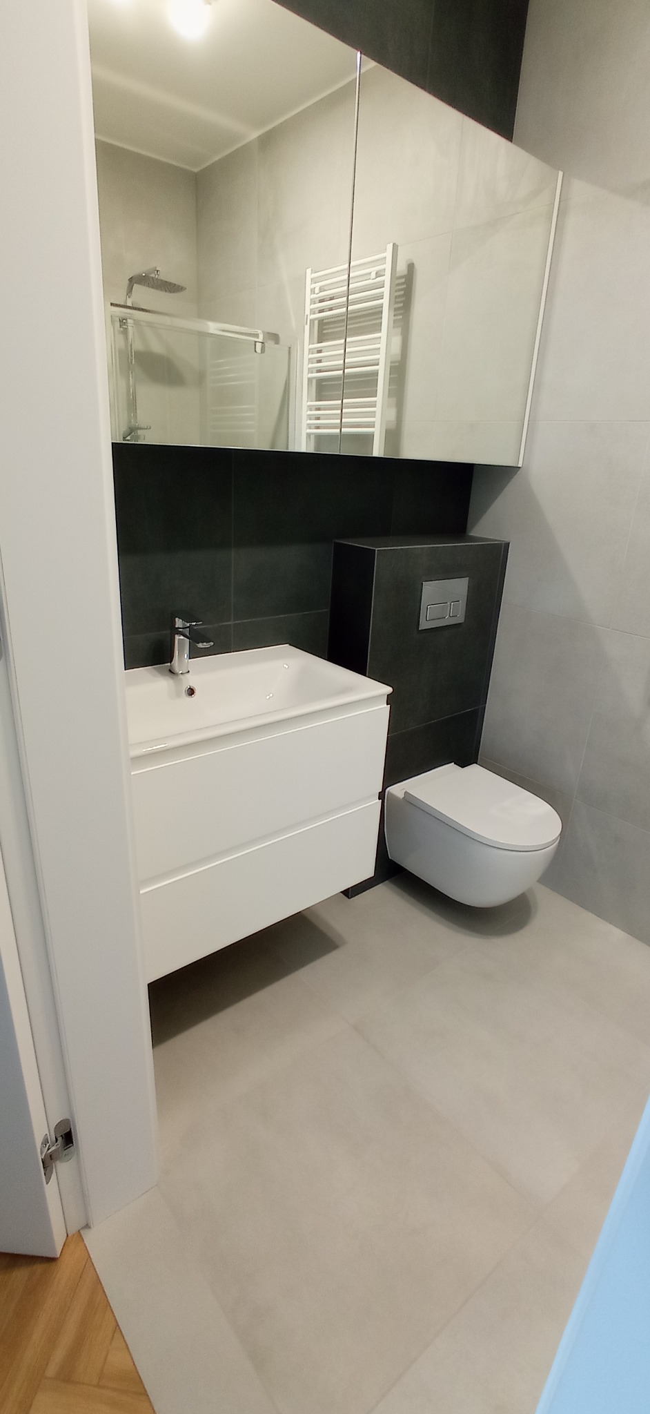 Nowoczesna łazienka w szarościach: biała umywalka z szafką, podwieszana toaleta z czarną obudową, duże lustro z odbiciem kabiny prysznicowej i grzejnika.