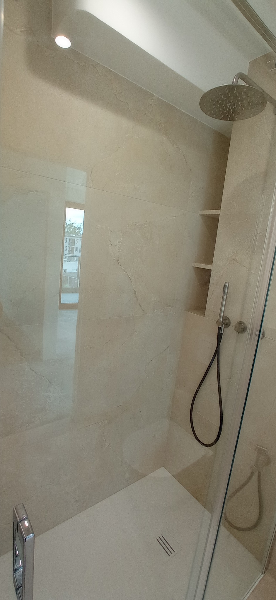 Beżowa kabina prysznicowa z deszczownicą i wnęką w ścianie, wykończona płytkami imitującymi marmur. Widoczne detale: bateria, odpływ liniowy i fragment szklanej ściany.