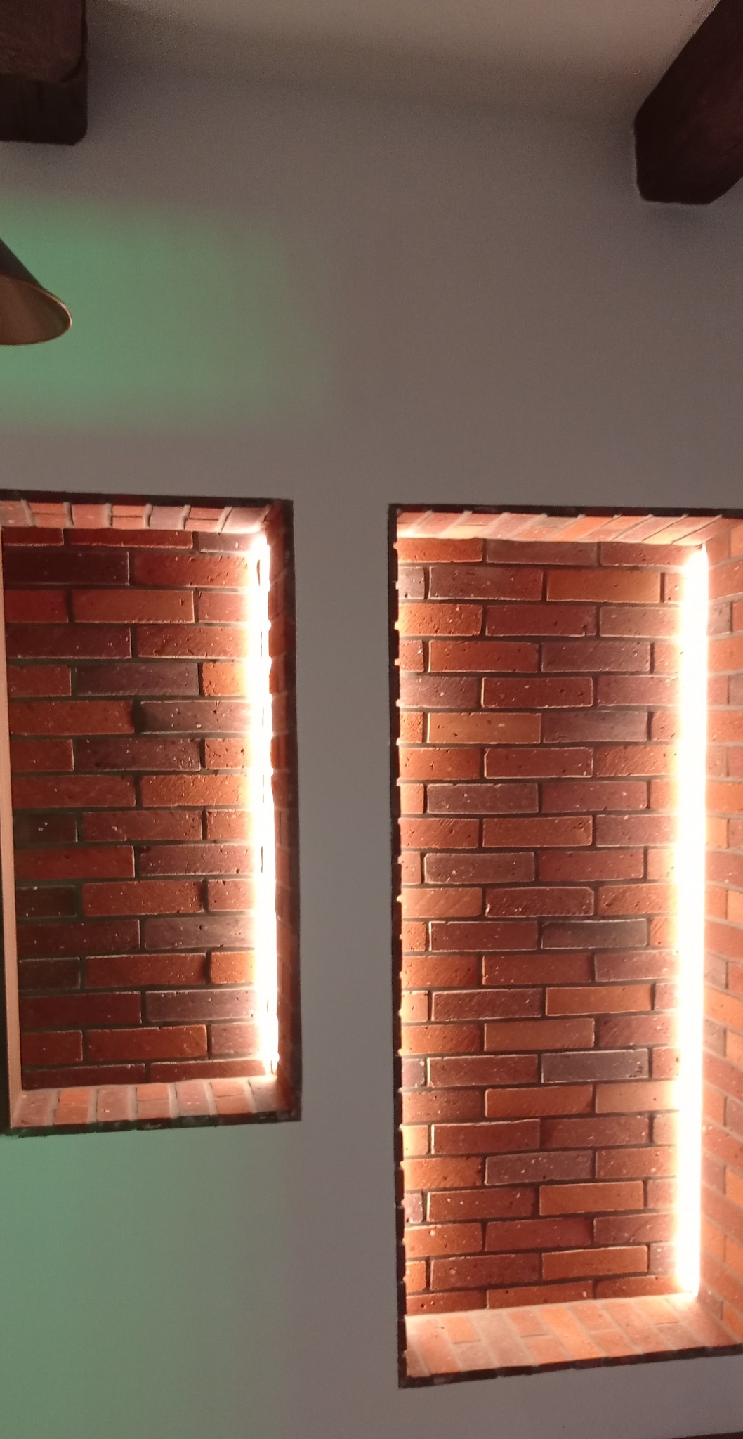 Dwa ceglane wnęki z podświetleniem LED w białej ścianie, tworzące dekoracyjny element wnętrza. Widoczne fragmenty drewnianego sufitu.