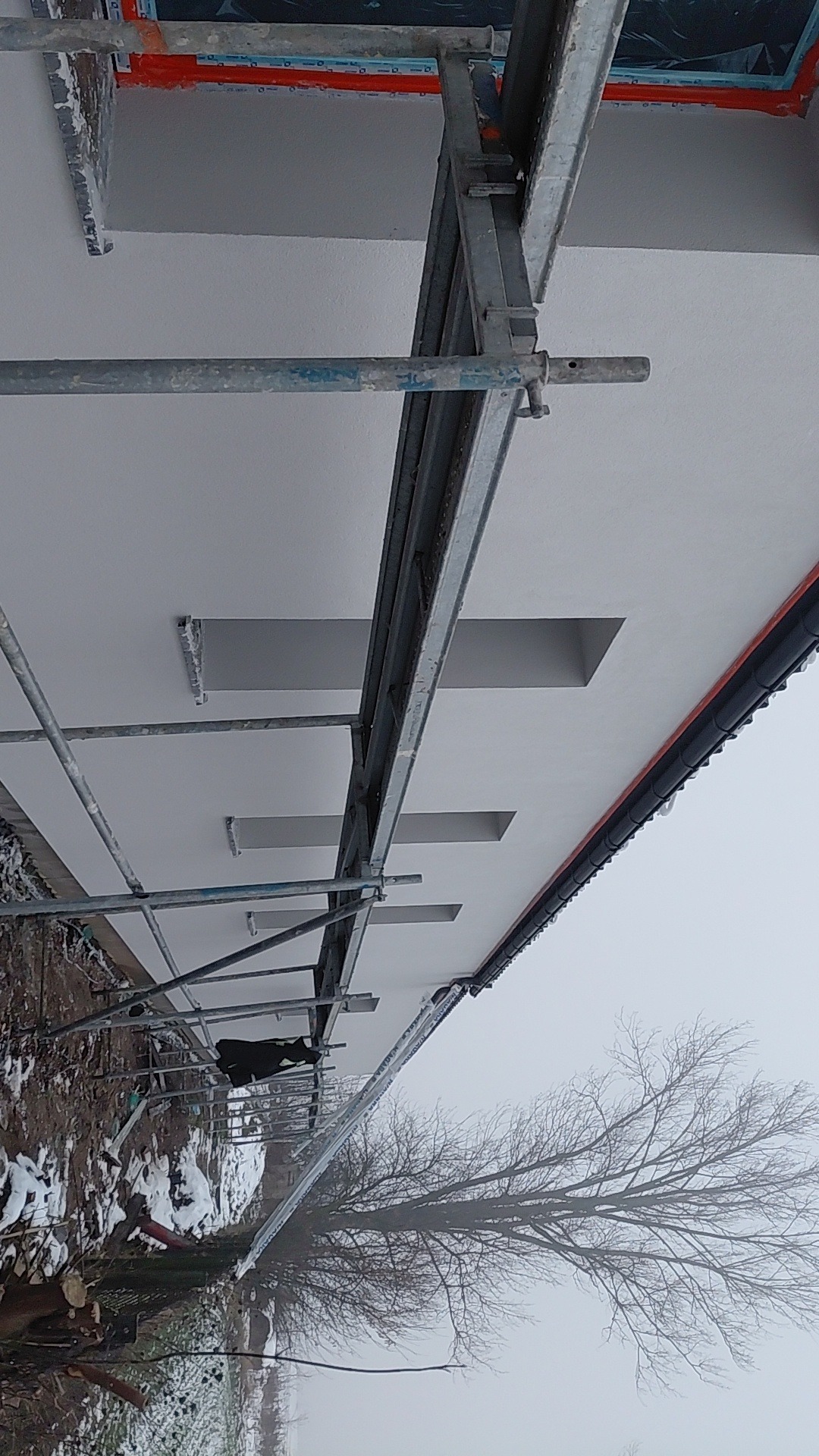 Świeżo otynkowana elewacja budynku z widocznymi otworami okiennymi, częściowo zasłonięta rusztowaniem. W tle drzewo bez liści i mglista pogoda.