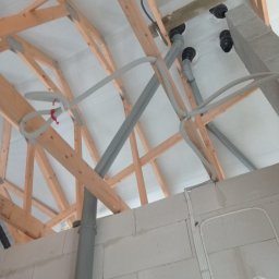 BRACIA HYDRAULICY ŁUKASZ PARJASZEWSKI - Szare rury wentylacyjne prowadzone pod drewnianą konstrukcją dachu, widoczne elementy instalacji w surowym stanie, ściany z białych bloczków.