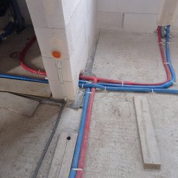 BRACIA HYDRAULICY ŁUKASZ PARJASZEWSKI - Wnętrze w trakcie prac hydraulicznych: czerwono-niebieskie rury prowadzone wzdłuż ściany z betonu komórkowego, widoczne elementy instalacji i podłoże w stanie surowym.