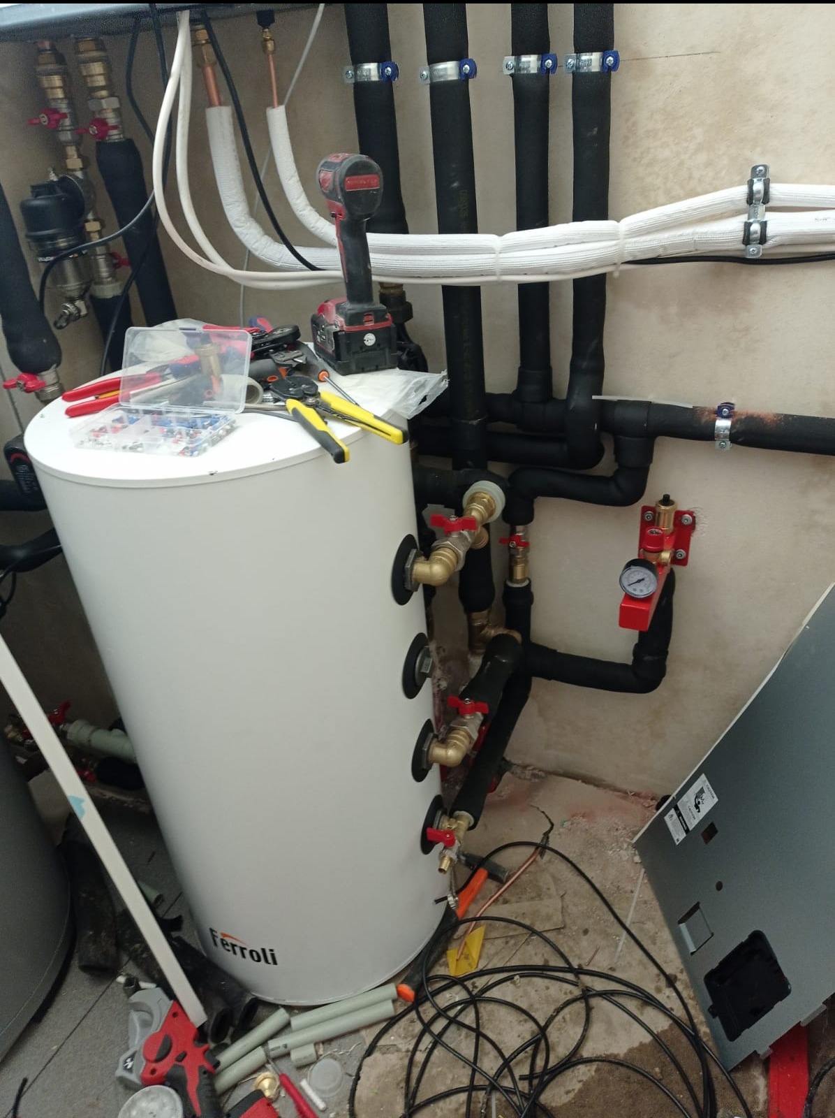 Biały zasobnik ciepłej wody użytkowej Ferroli z podłączonymi rurami i zaworami, narzędzia pracy na górze, instalacja w trakcie montażu.