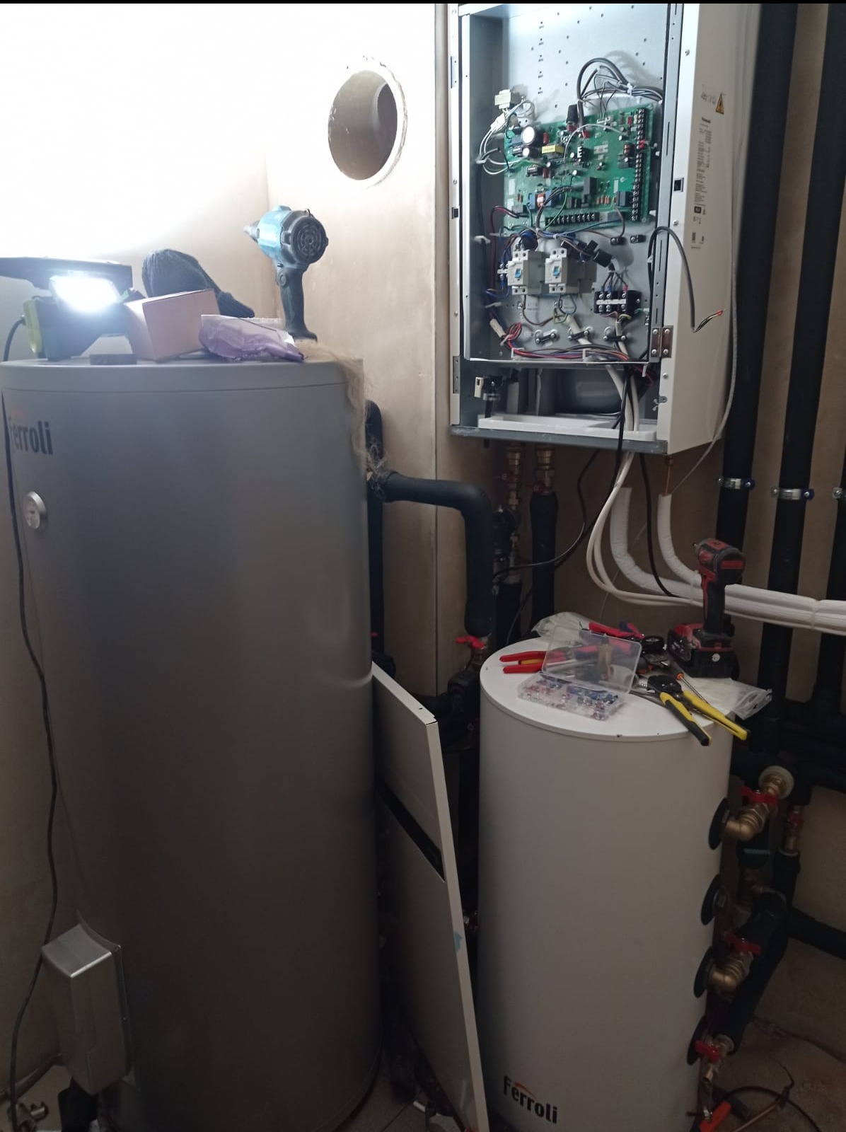 Dwa zbiorniki Ferroli i otwarty piec gazowy z widoczną elektroniką, narzędzia na zbiorniku, rury instalacji grzewczej przy ścianie. Instalacja w trakcie montażu.