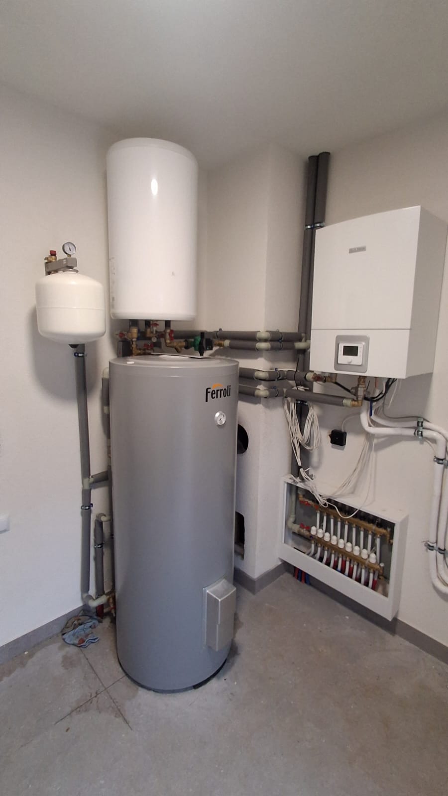 Szary bojler Ferroli z białym zbiornikiem wyrównawczym, piec gazowy i rozdzielacz ogrzewania podłogowego w kotłowni. Widoczne rury i instalacje.