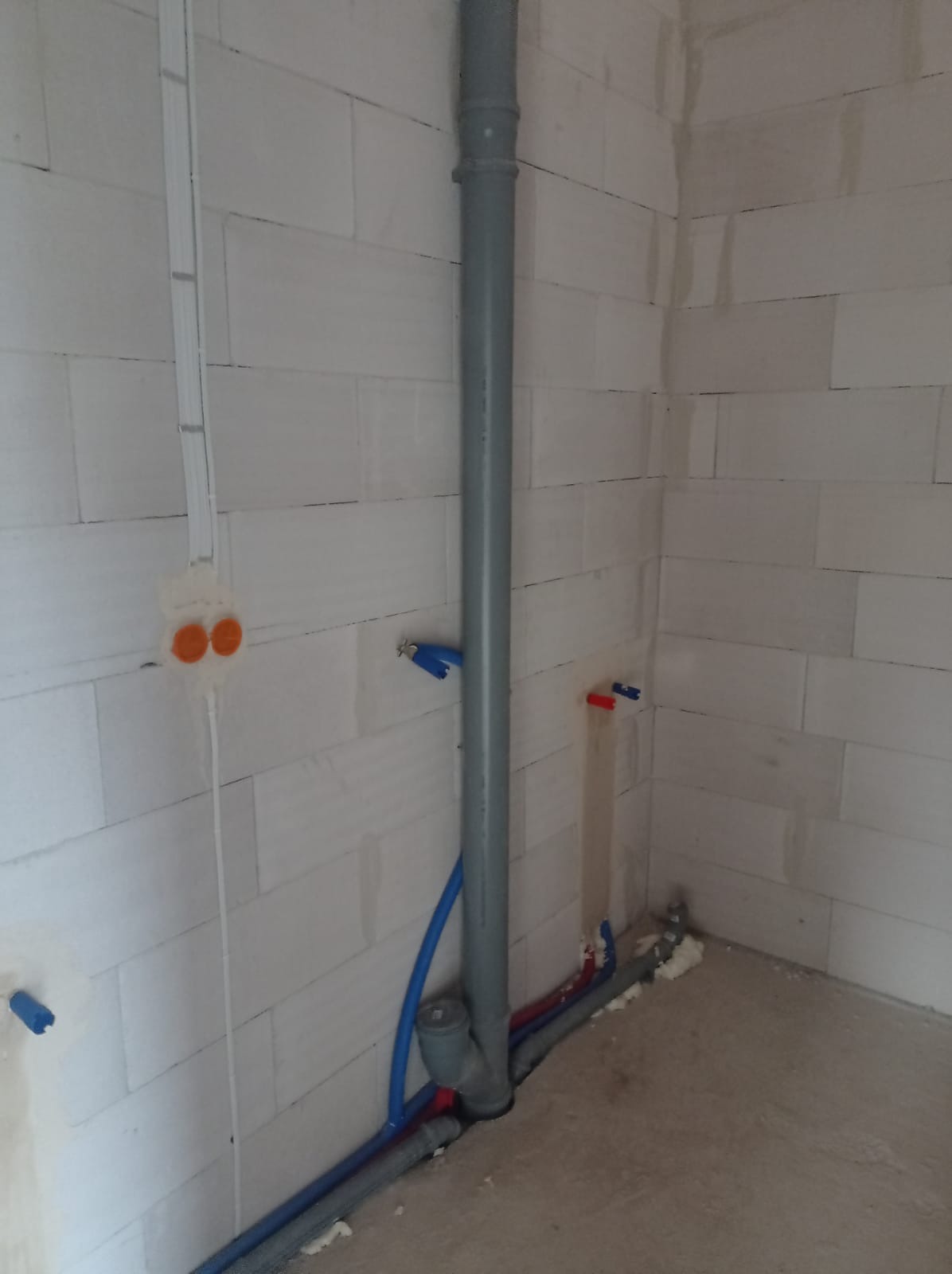 Szare rury kanalizacyjne i niebieskie rury wodne w rogu pomieszczenia z białych bloczków, instalacja w trakcie montażu, widoczne elementy mocujące.