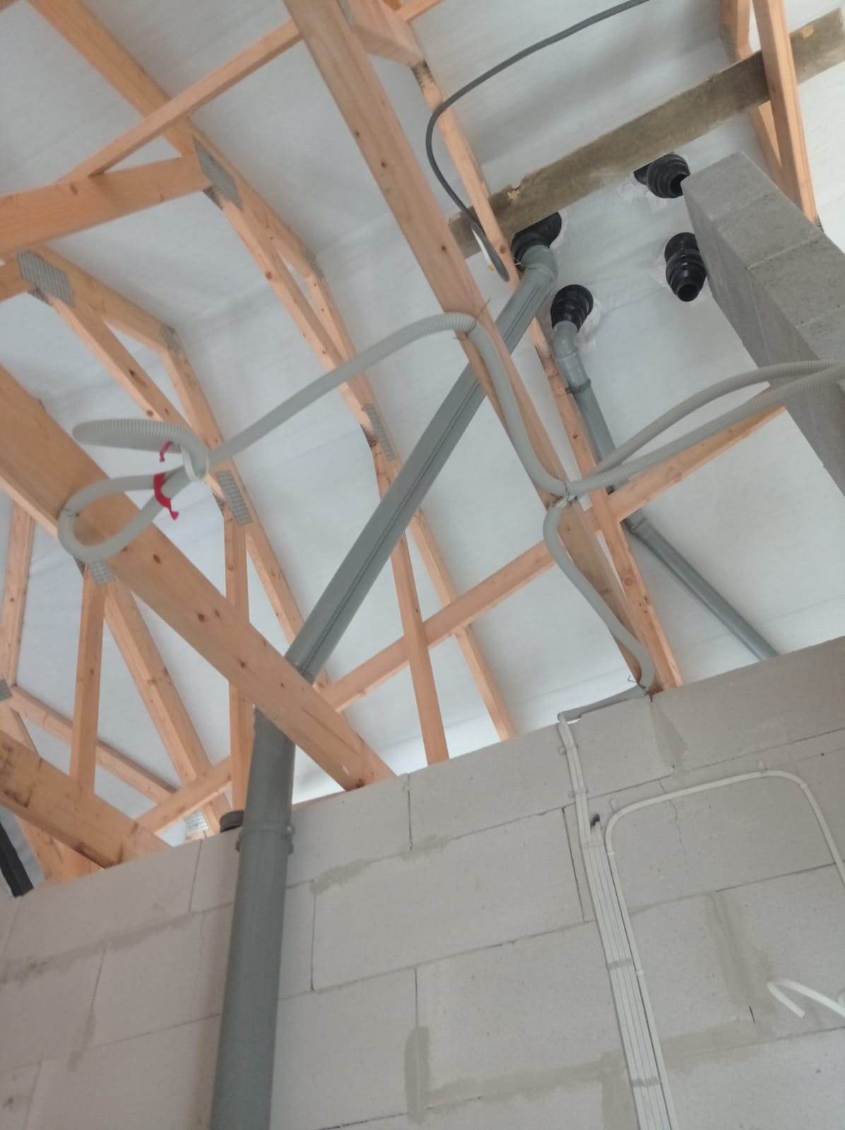 Szare rury wentylacyjne prowadzone pod drewnianą konstrukcją dachu, widoczne elementy instalacji w surowym stanie, ściany z białych bloczków.