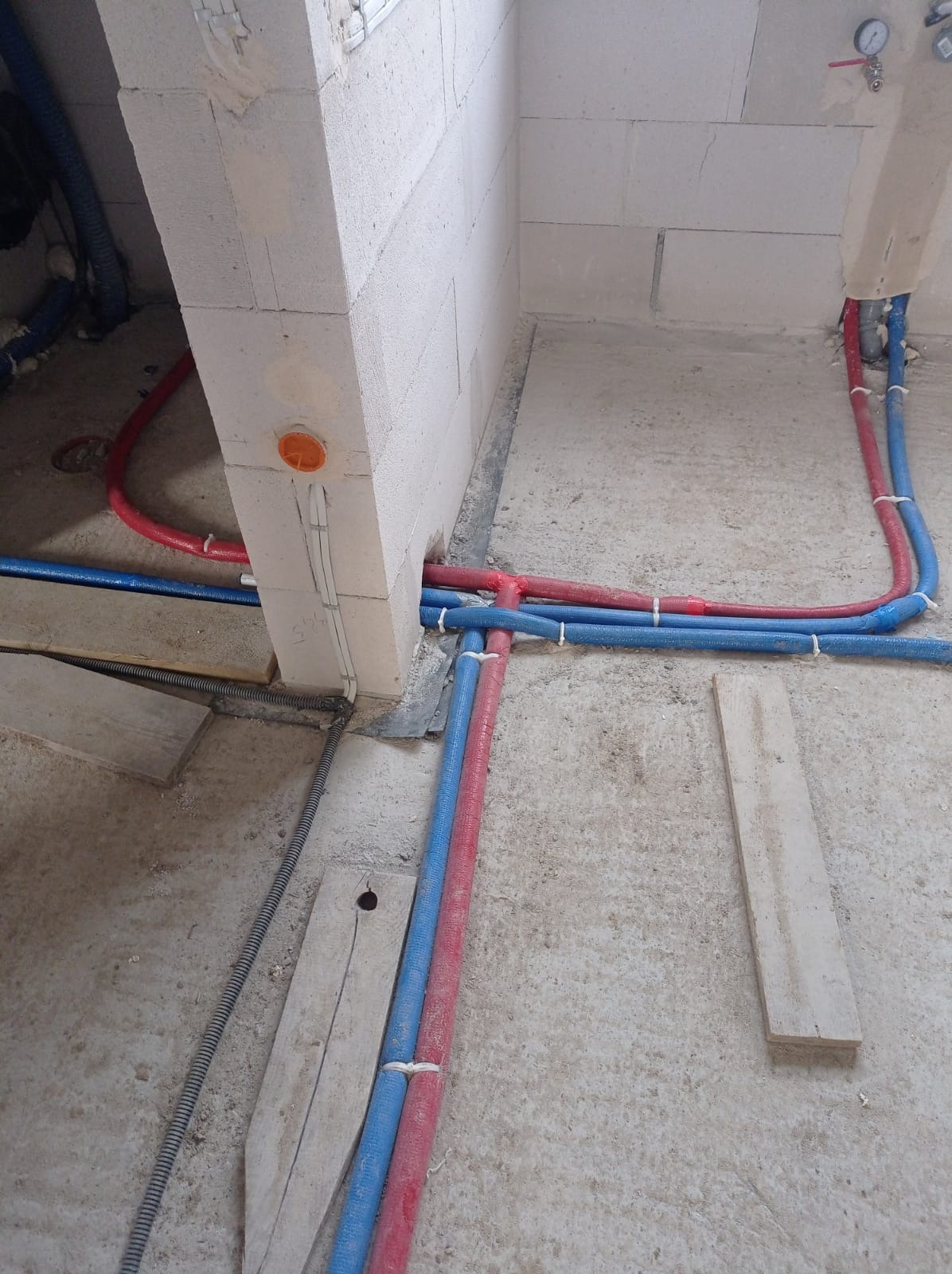 Wnętrze w trakcie prac hydraulicznych: czerwono-niebieskie rury prowadzone wzdłuż ściany z betonu komórkowego, widoczne elementy instalacji i podłoże w stanie surowym.