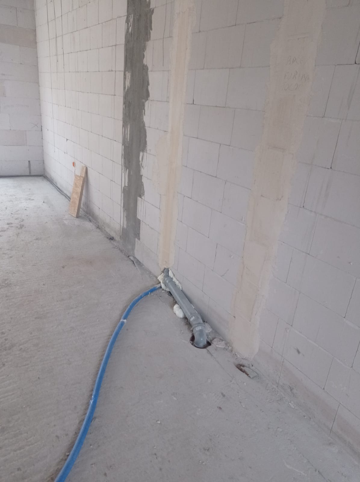 Szara rura kanalizacyjna wystaje z podłogi w surowym wnętrzu, zabezpieczona pianką montażową. Niebieski wąż leży na szarym betonie. Ściany z białych bloczków.
