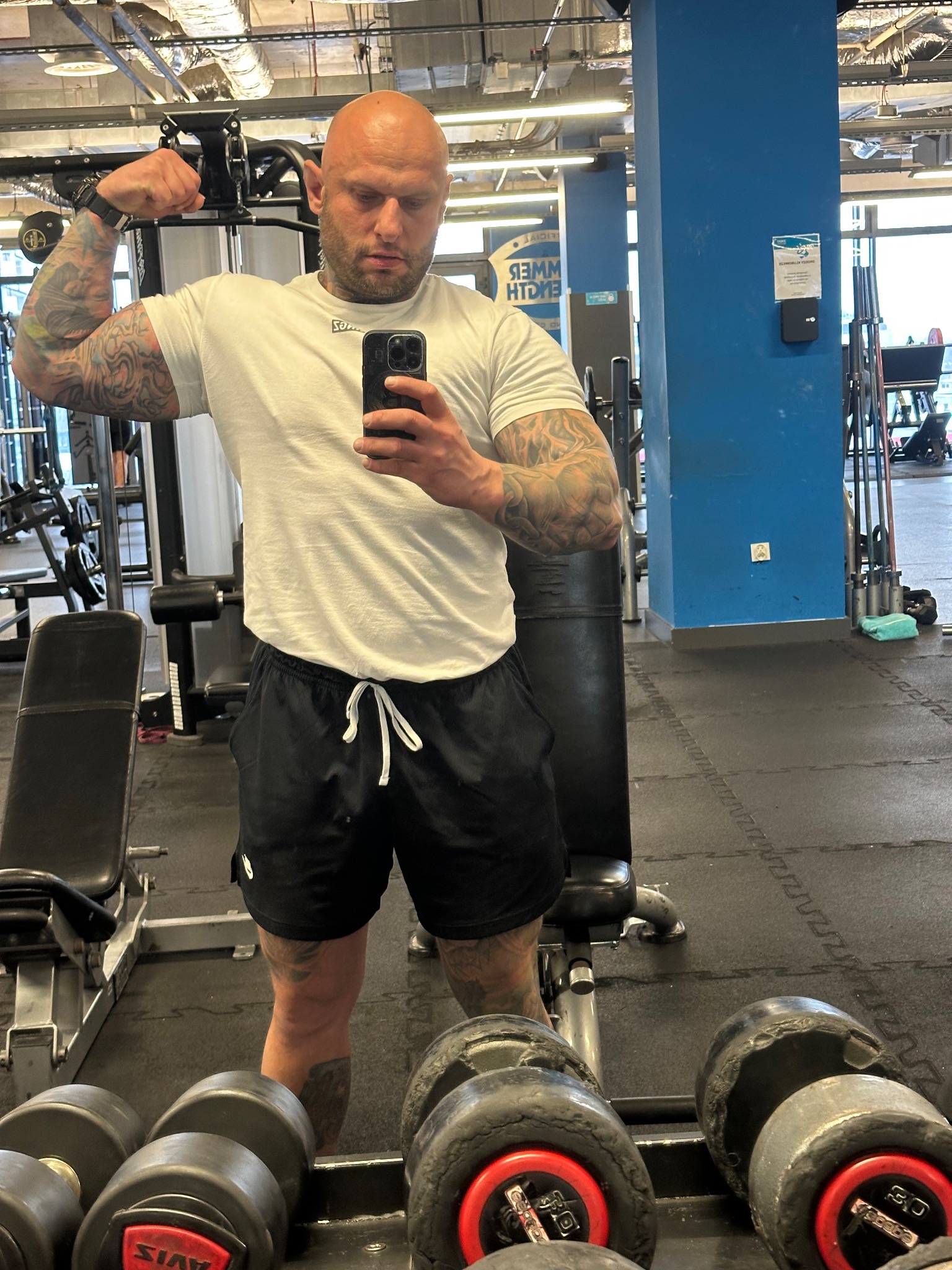Mężczyzna z tatuażami prezentuje bicepsy, robiąc selfie w siłowni. W tle widoczne sprzęty do ćwiczeń i kolumna w kolorze niebieskim.