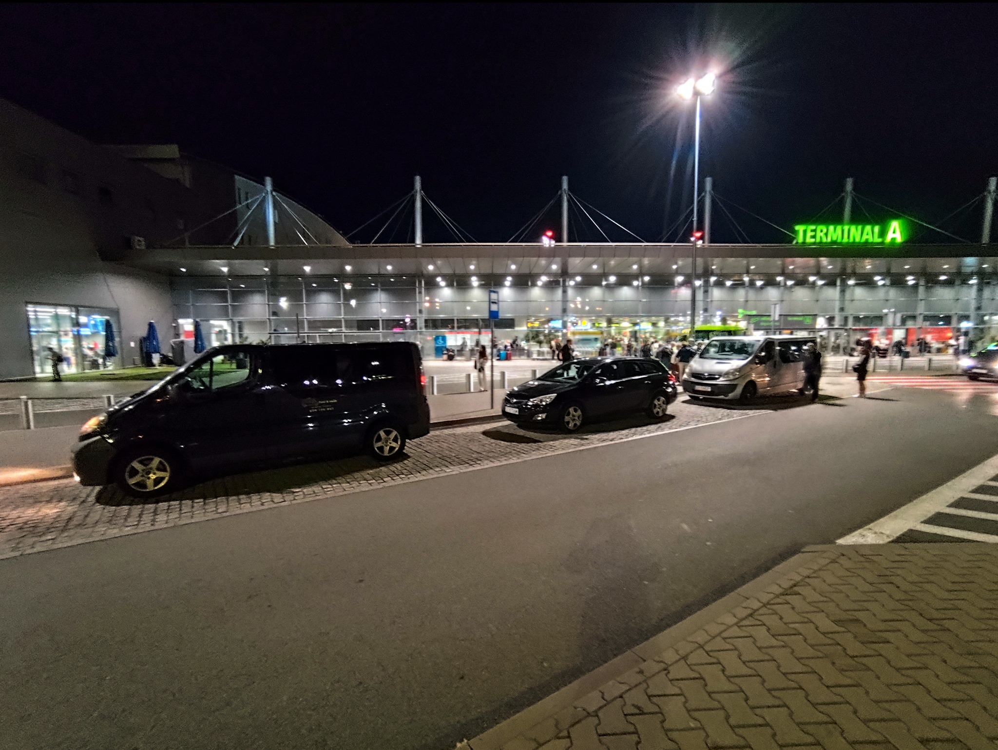 Nocny widok na Terminal A lotniska z zaparkowanymi busami i samochodami osobowymi, oczekującymi na pasażerów. Oświetlony budynek terminala w tle.