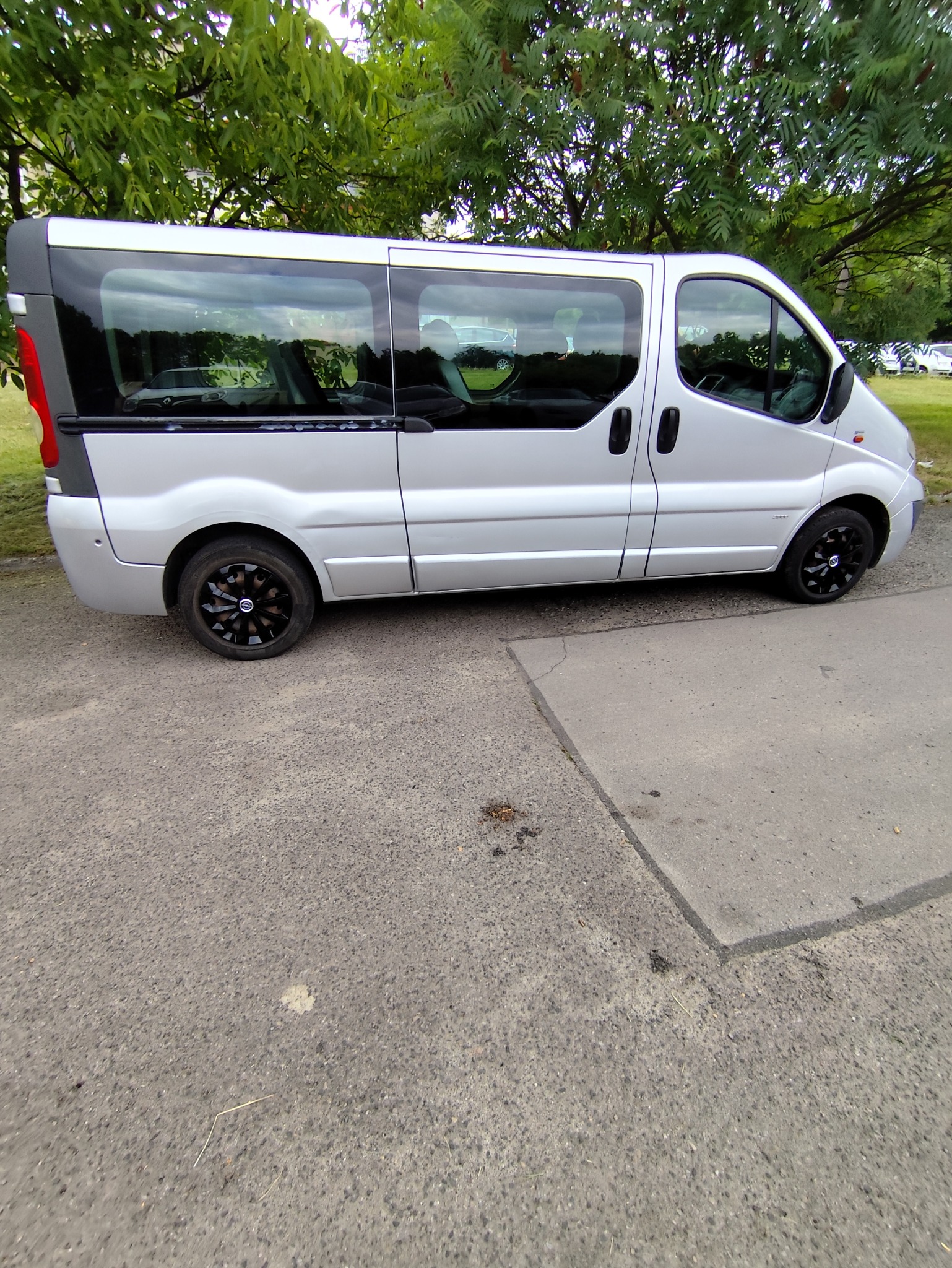 Srebrny bus z czarnymi felgami zaparkowany na betonowym podłożu, z zielonymi drzewami w tle. Widok z boku, lekko z dołu. Szyby odbijają otoczenie.