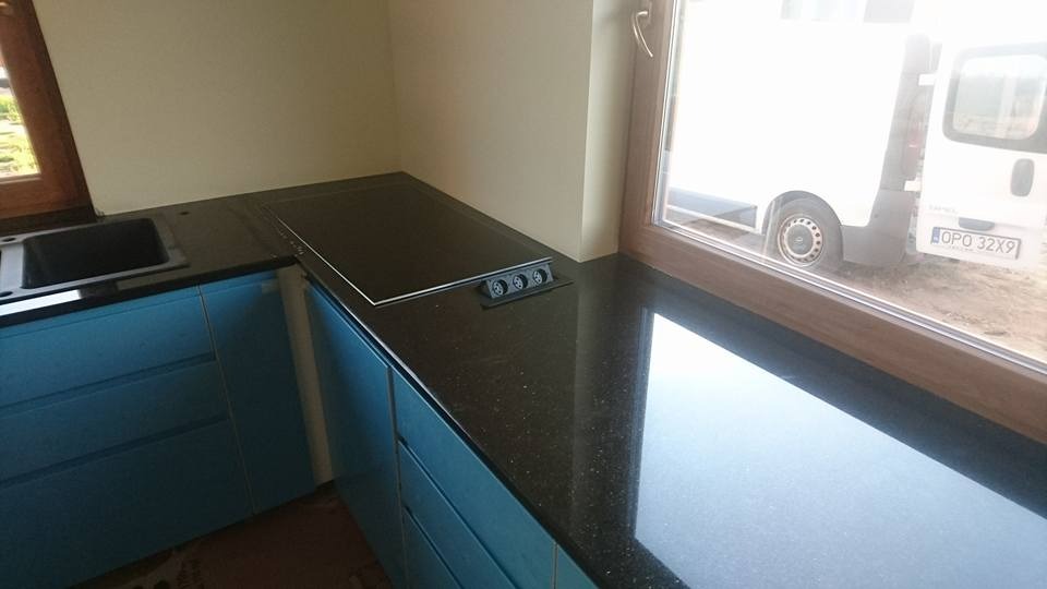 Błyszczący, czarny blat kamienny z wbudowaną płytą grzewczą i gniazdkami, zamontowany w kuchni z błękitnymi szafkami. Widok z okna na zewnątrz.