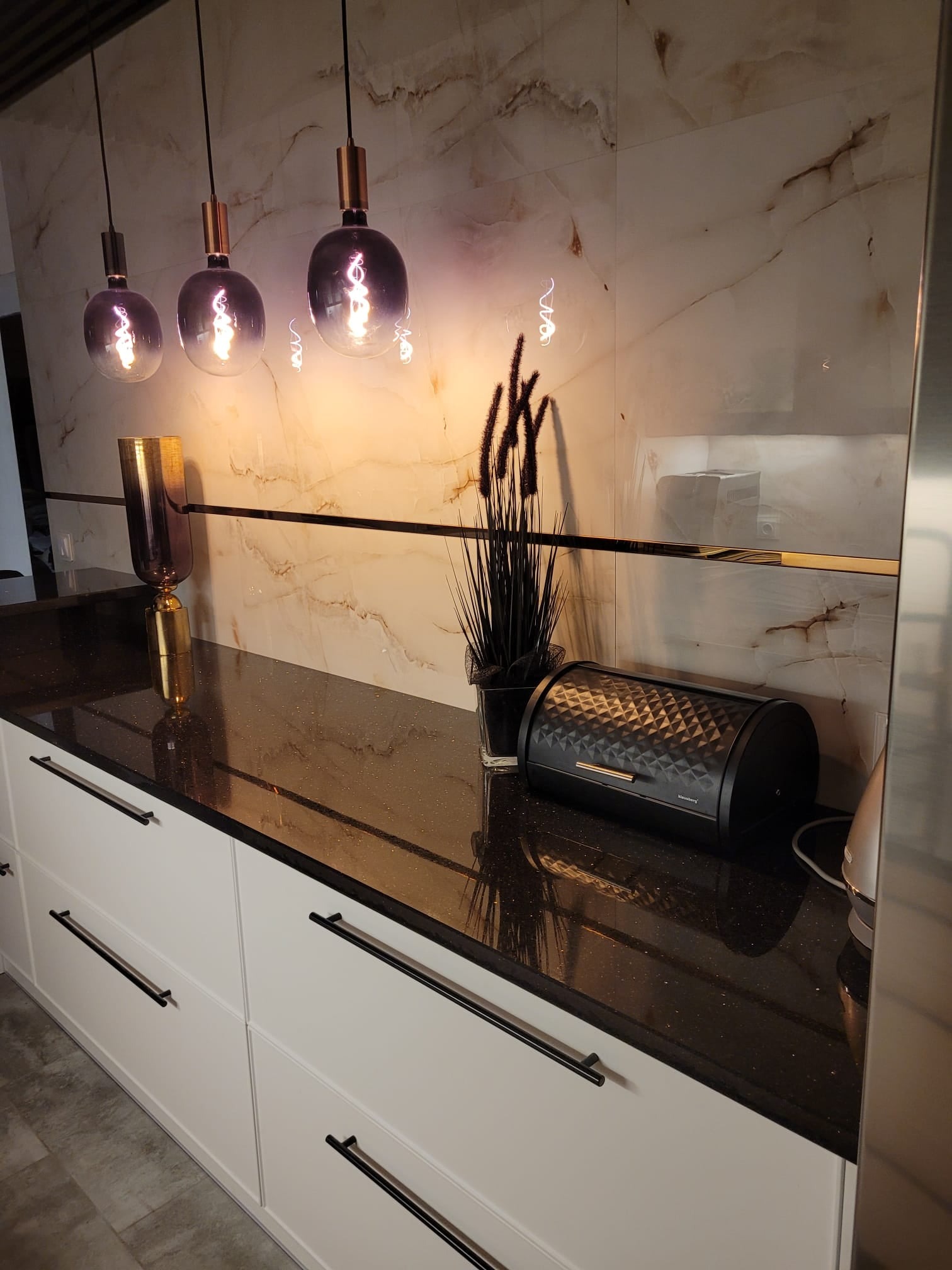 Błyszczący, czarny blat z konglomeratu w nowoczesnej kuchni, oświetlony trzema wiszącymi lampami. Widoczne białe szafki z długimi, czarnymi uchwytami i marmurowa ściana.