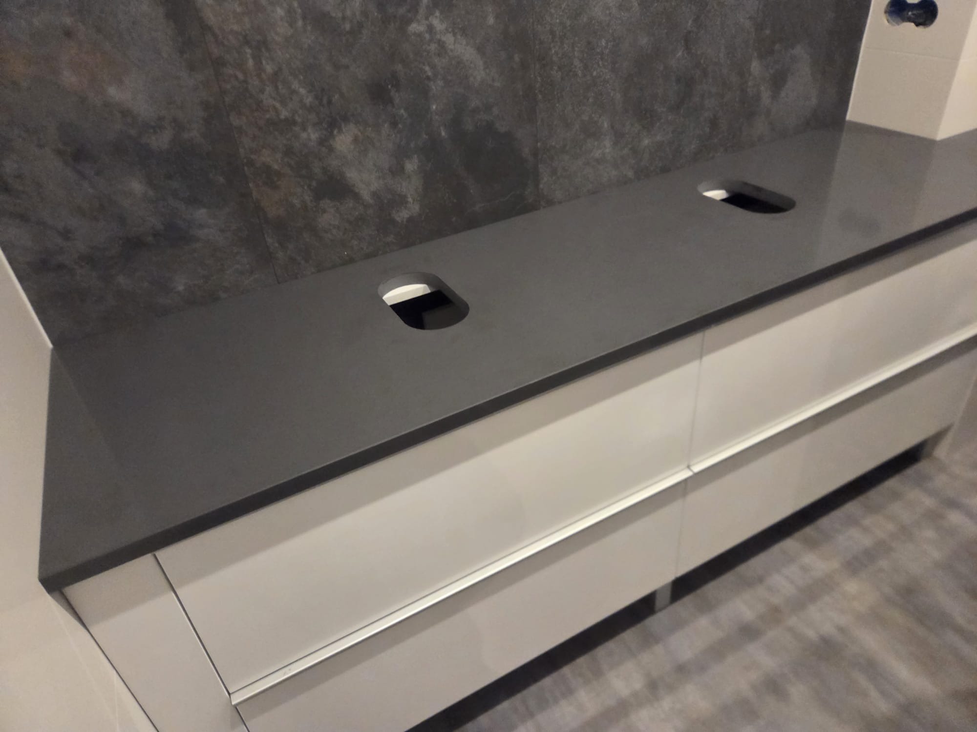 Szary blat kamienny z dwoma otworami na umywalki, zamontowany na białych szafkach w łazience z szarą ścianą. Ujęcie z perspektywy.