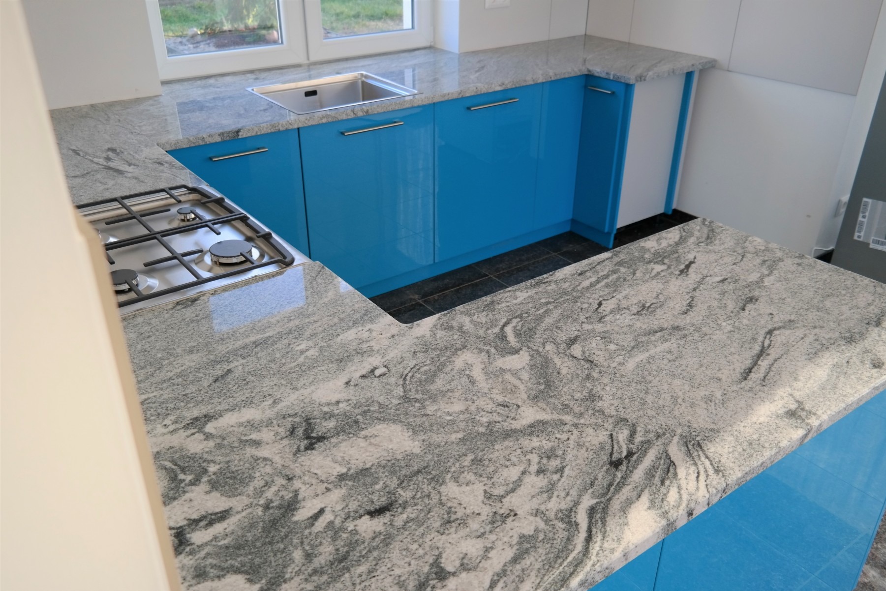 Jasnoszary blat kamienny z kuchenką gazową i zlewozmywakiem, zamontowany na niebieskich szafkach kuchennych. Widok z góry, oświetlenie naturalne.