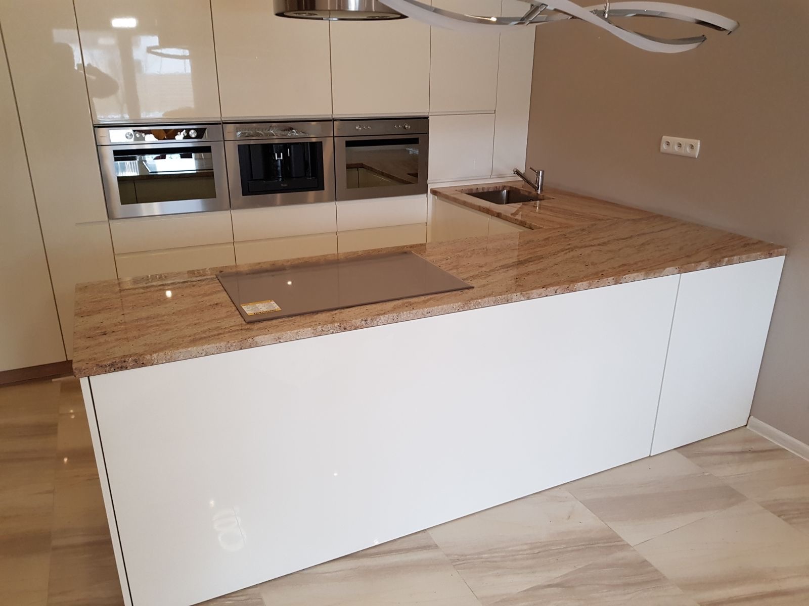 Beżowy blat kamienny z wbudowaną płytą grzewczą i zlewem w nowoczesnej, jasnej kuchni z białymi szafkami. Widoczne gniazdka elektryczne na ścianie.