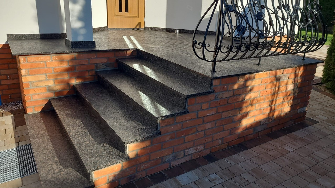 Zewnętrzne schody z ciemnego kamienia, wykończone cegłą, prowadzące do drzwi wejściowych. Balustrada z kutego żelaza. Widoczne promienie słoneczne.